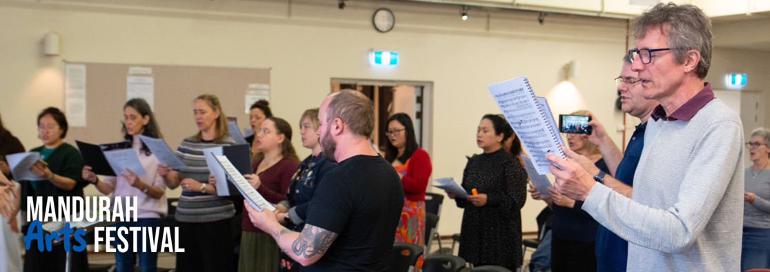 A group of people stand in a room reading sheet music and singing together